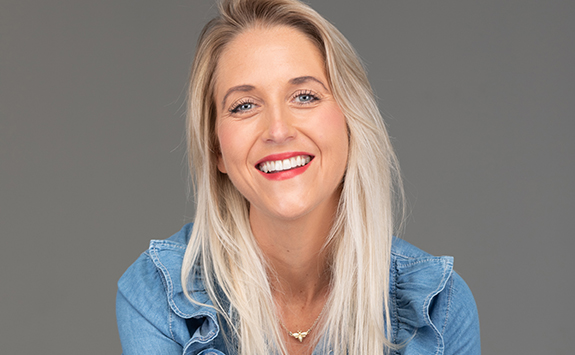 A smiling woman with blonde hair and wearing a denim shirt sits in front of a grey background